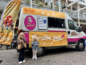 Twee jonge meisjes eten een schepijsje en staan voor een ijscokar voor een banner waar 'gratis ijsje' opstaat.